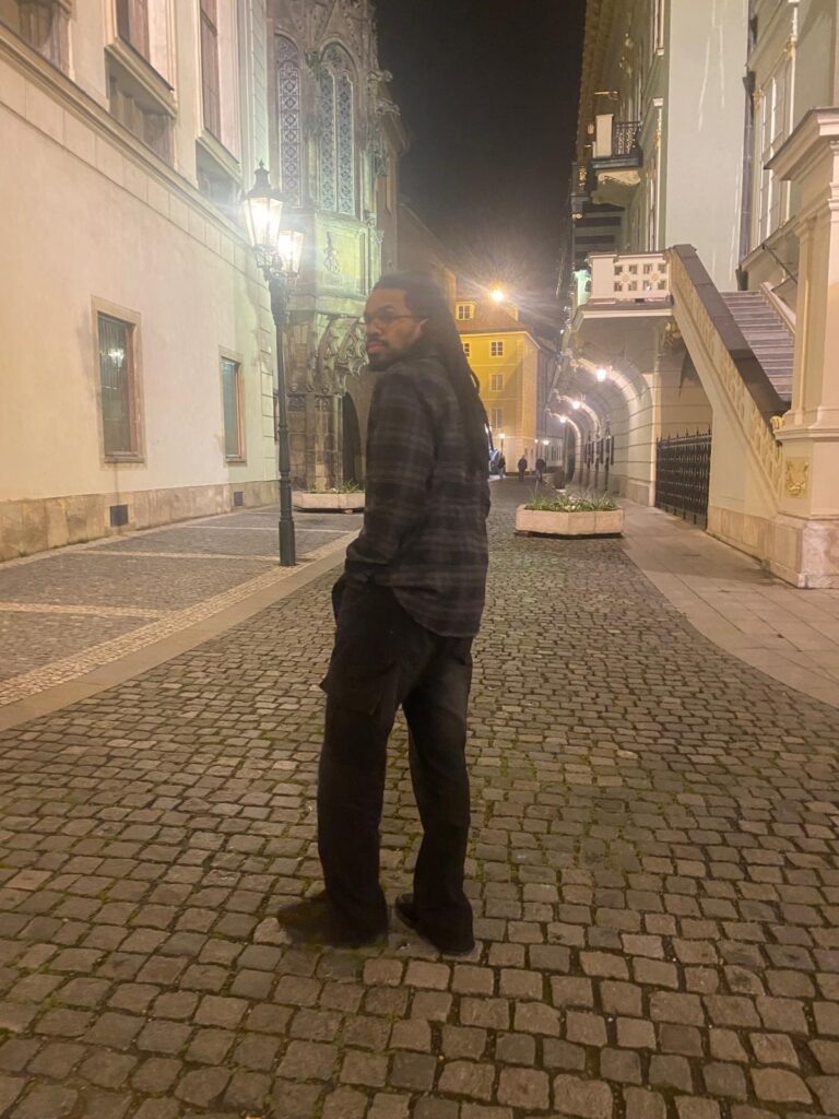 Javan C., author of The Creary Post, stands on a cobblestone street at night, glancing back at the camera. The street is illuminated by vintage lamps, with historic buildings featuring intricate architecture in the background.