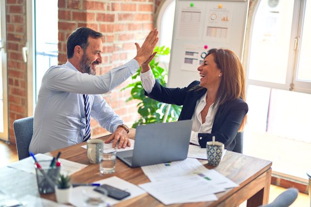 Two cybersecurity professionals celebrating a successful project in an office with documents, charts, and a laptop on the table.