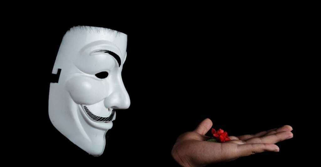 Side view of an anonymous mask facing a hand holding a red flower against a black background.