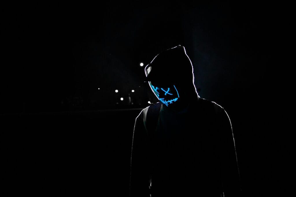 Silhouette of a person in a hoodie wearing a glowing LED mask against a dark background.